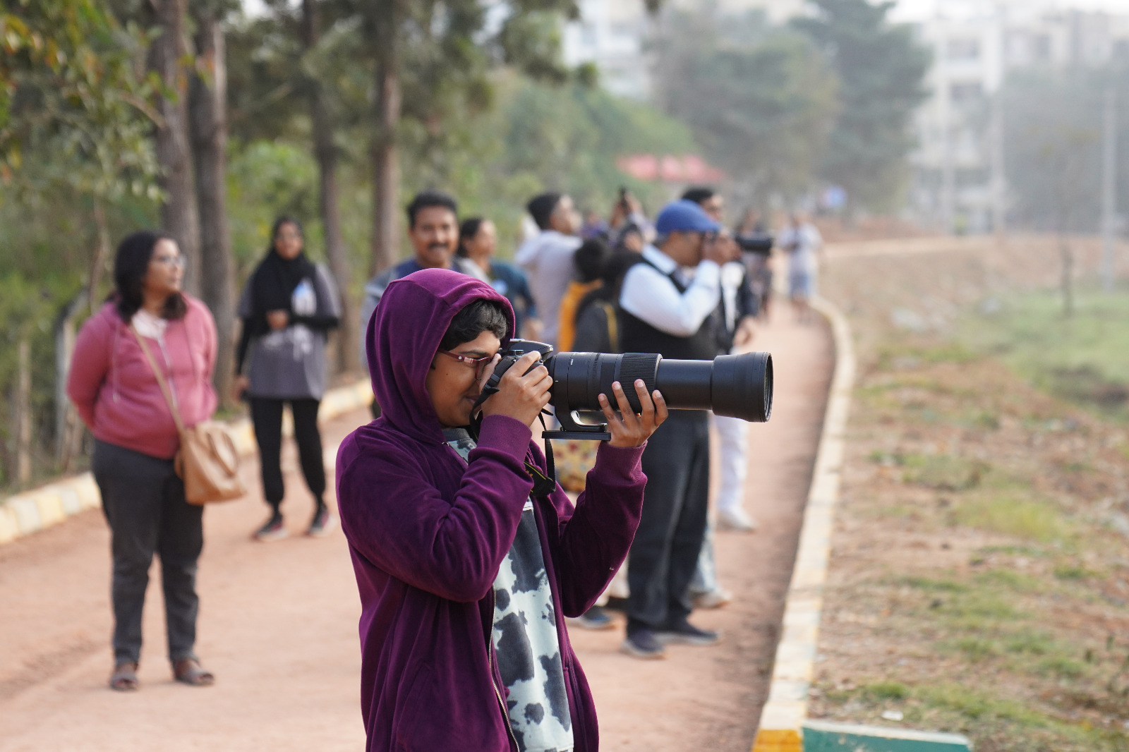 Nature walk & Birding A boy wearing a purple jacket, and holding a camera in the foreground, with a group of people looking towards a lake in the background.