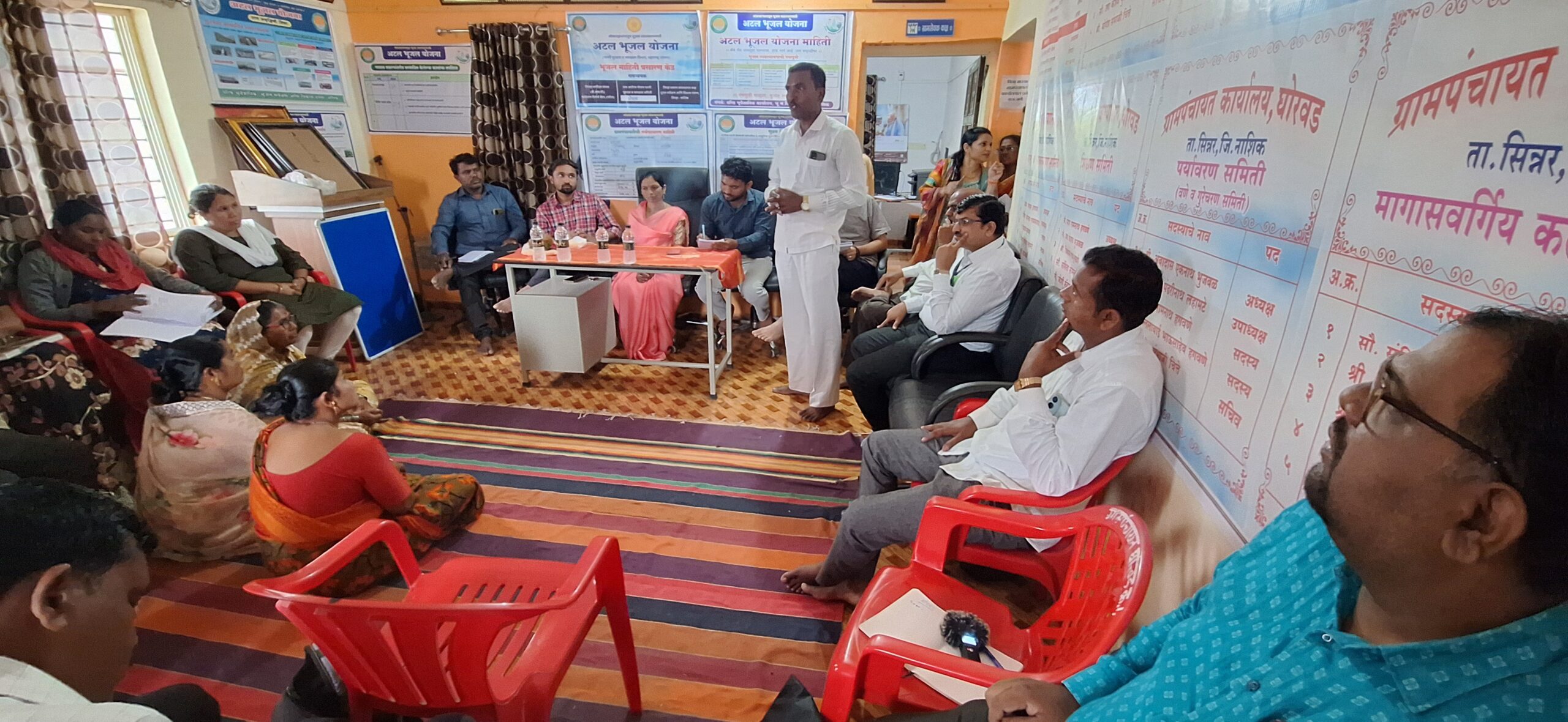 Multiple people stand and sit on the floor of a small building. The floor is covered with red-hued carpets and some red plastic chairs. The walls have posters in Devanagari script. 