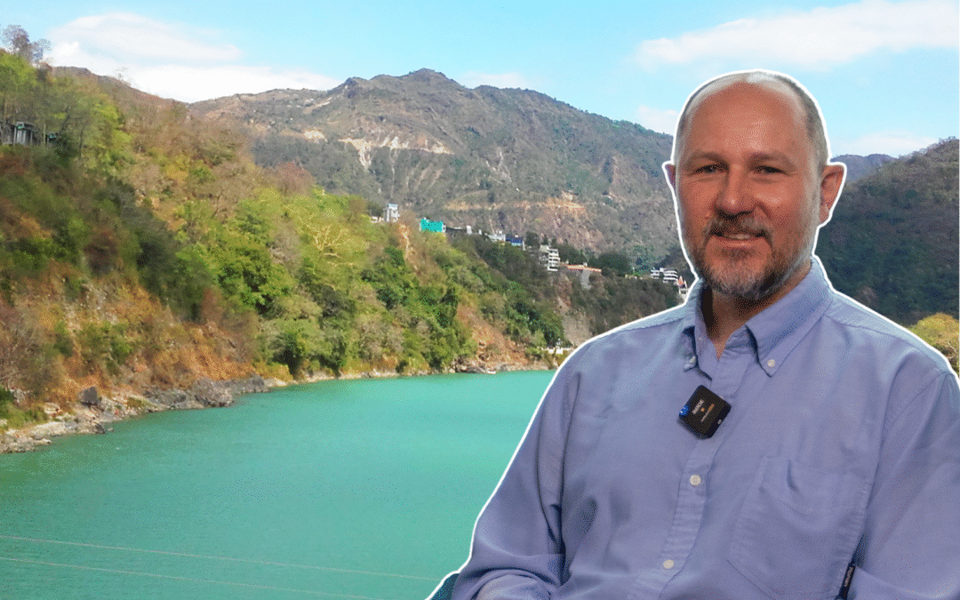 Image of Alan MacDonald in the foreground, and an image of the Ganges river basin in the background. Clear water flows in the river from hills, towards the viewer.