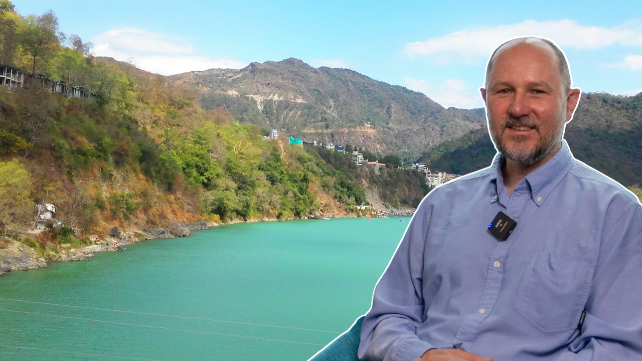 Image of Alan MacDonald in the foreground, and an image of the Ganges river basin in the background. Clear water flows in the river from hills, towards the viewer.