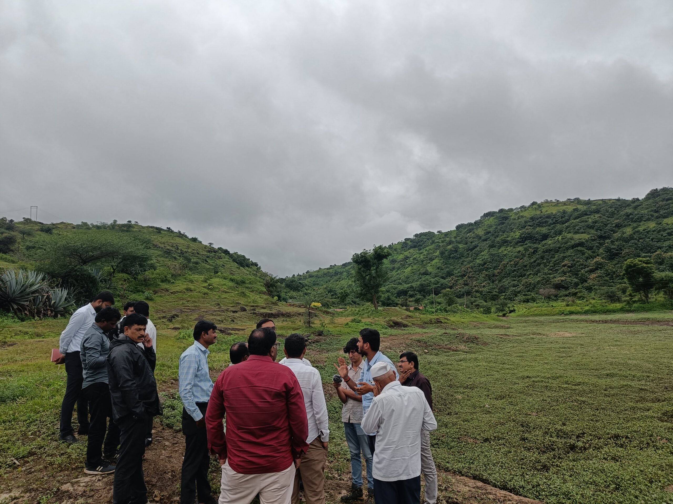 Image with a green valley in the background and a group of people speaking to each other in a huddle in the foreground. The sky is cloudy. 