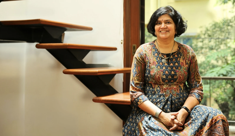 Image of Veena Srinivasan, sitting on wooden stairs within a room.