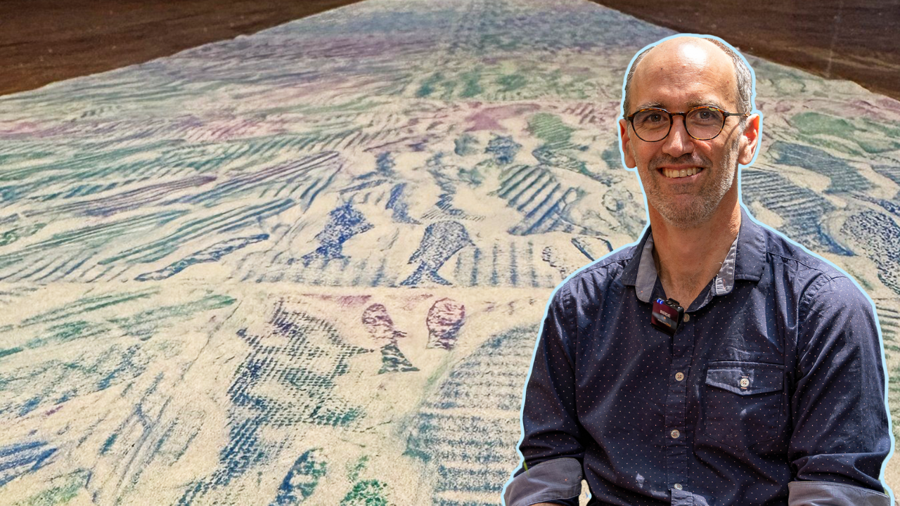 A man wearing glasses and a blue shirt in the foreground, a long sheet of brown paper with fish drawn on it in the background. 