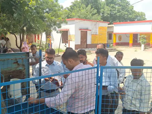A group of men inspecting a device in the foreground, with a series of small, white building structures in the background.