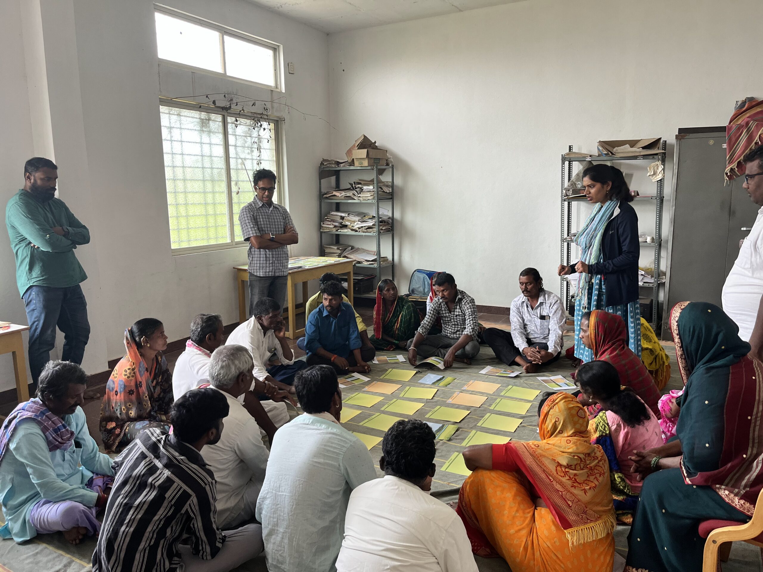 There are a few people standing and multiple people sitting in a circle inside a room. In the middle of the circle are sheafs of paper arranged in a rectangular formation.